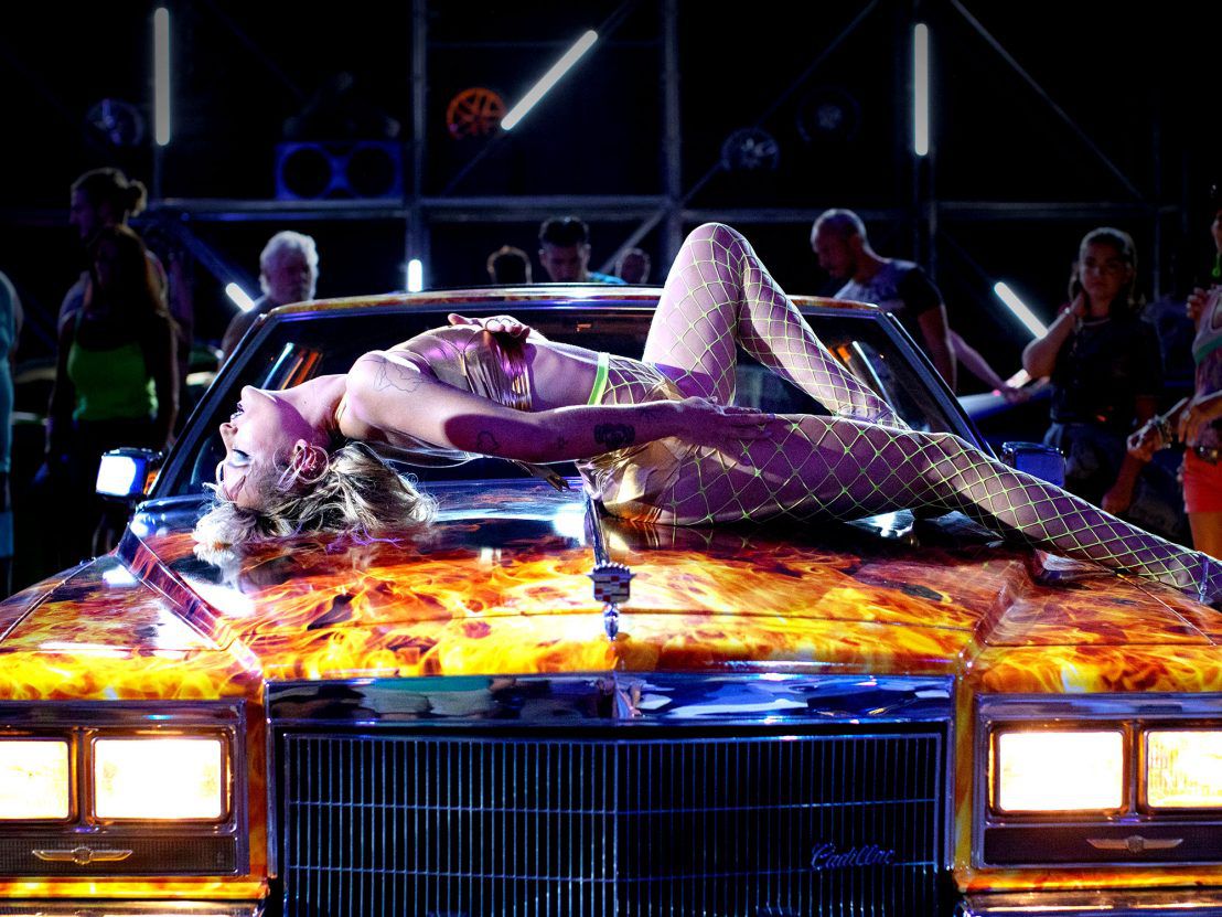 A young woman straddles atop a neon-painted car hood in Titane